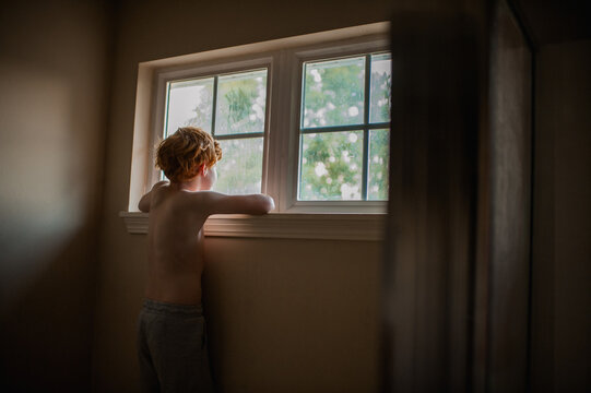 Young shirtless boy peacefully stares out soft lit windows