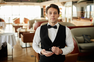 Portrait of young Caucasian man wearing formal uniform standing in restaurant holding notepad looking into camera working as waiter in dining area