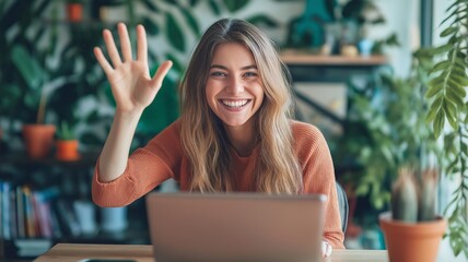 Young woman waves hello while working on laptop in a bright space filled with plants
