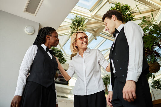 Caucasian middle aged woman greeting Black young adult woman and Caucasian young adult man in restaurant uniform, smiling and interacting in modern restaurant setting