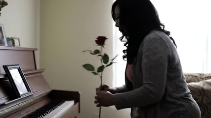 A woman holds a single red rose, standing by a piano with family photos, appearing thoughtful and reflective