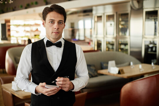 Portrait of young Caucasian man standing in restaurant holding notepad and pen wearing formal uniform looking into camera working as waiter