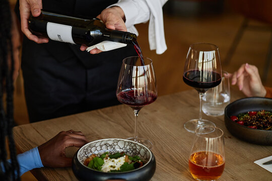 Man pouring red wine into glass for diverse group of young adults sitting at restaurant table with food and drinks - Powered by Adobe