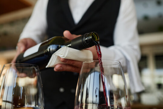 Man pouring red wine into glasses, wearing formal vest and shirt, hands visible holding bottle and napkin, restaurant worker serving drinks to customers
