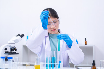 A young Asian female scientist in lab coat and gloves holds a glass pipette carefully in a science...