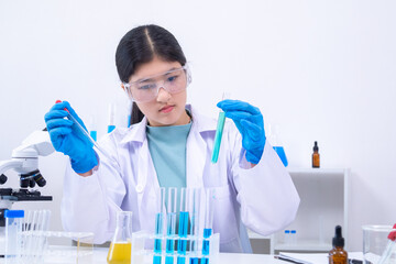 A young Asian female scientist in lab coat and gloves holds a glass pipette carefully in a science...
