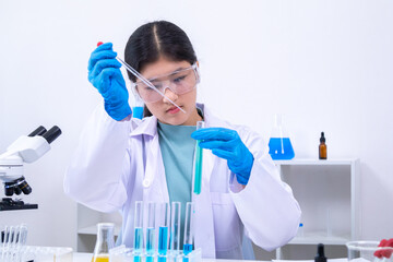 A young Asian female scientist in lab coat and gloves holds a glass pipette carefully in a science...