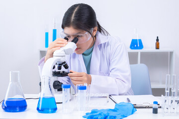 A young Asian female scientist in lab coat observes samples through a microscope in a clean science...