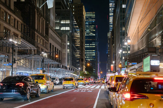 Yellow taxis and a black car drive through Midtown Manhattan at night under skyscrapers and streetlights. The scene features busy traffic, scaffolding, and the bright glow of the city.