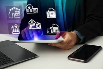 Real estate agent looking for housing via tablet at white table, closeup. Illustrations of different buildings over device
