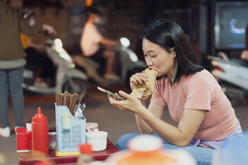 Woman enjoying street food while using her smartphone in a bustling Hanoi night market