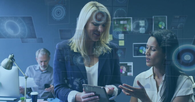 Pointing and gesturing, two female colleagues examining tablet at office desk, with sticky notes