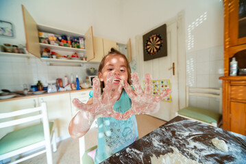 Little girl making dough in messy kitchen, showing floury hands