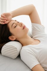 Obraz premium A soft and clean studio photo of a young woman lying on her back with her eyes closed, resting her head on a striped cylindrical neck pillow. 