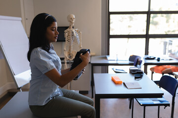 Healthcare trainee inspecting VR headset at bench with skeleton and books by windows