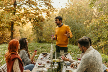 Friends enjoying thanksgiving dinner outdoors in beautiful autumn forest setting as man gives toast