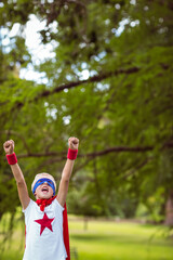 Boy wearing homemade superhero costume and raising fists in sunlit park under tree branches
