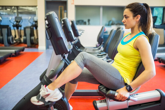 Fototapeta Woman pedaling recumbent bike in fitness studio, mirrored wall reflecting cardio machines