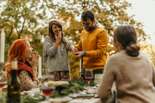 Man putting engagement ring on woman's finger during a surprise proposal at a garden party