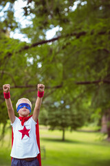 Child wearing red cape and blue mask raising fists in grassy park under tall trees
