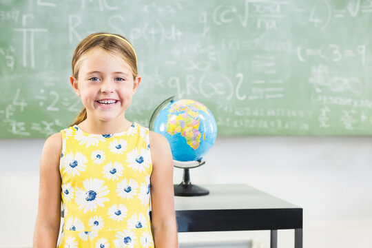 Girl standing in classroom in front of chalkboard with math formulas near desk with colorful globe