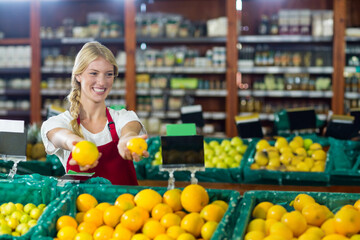 Female produce clerk wearing red apron holding two oranges behind green bins in produce section