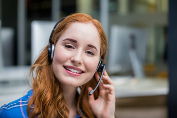 Red-haired support rep using headset and blue plaid shirt smiling at open-plan office environment