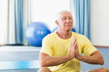 Senior man meditating on yoga mat in gym with stability balls while wearing yellow t-shirt