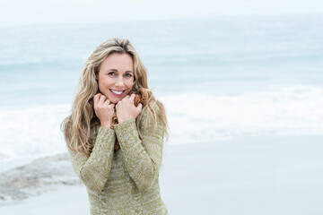 Woman standing on sandy beach in green sweater hugging teddy bear and smiling under overcast sky