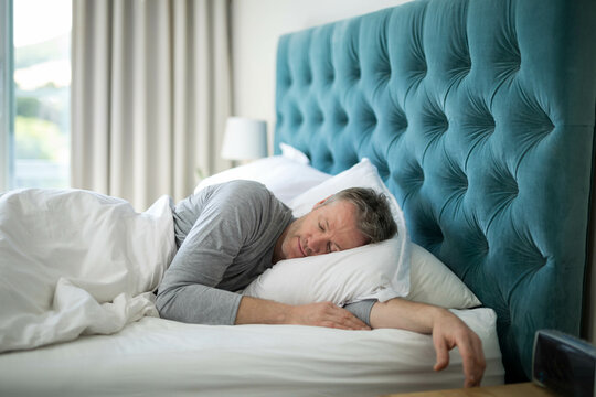 Mature man sleeping on side in master bedroom on white pillow under duvet, copy space