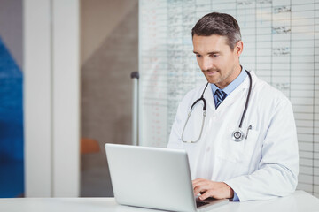 Doctor typing on silver laptop at desk with stethoscope and schedule board showing data behind him