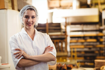 Female pastry chef standing in bakery wearing chef coat, hairnet amid racks, trays, copy space