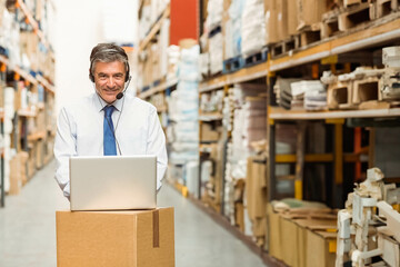 Warehouse manager operating open laptop on cardboard box in warehouse aisle with headset microphone