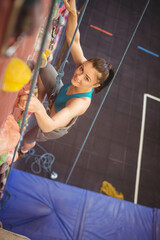 Female climber ascending artificial wall in climbing gym using rope, harness and holds, copy space