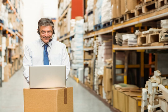 Warehouse manager operating open laptop on cardboard box in warehouse aisle with headset microphone - Powered by Adobe
