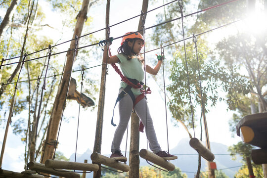 Child wearing orange helmet and red harness navigating suspended log rope course among sunlit trees - Powered by Adobe