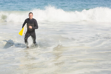 Middle aged man in black wetsuit emerging from ocean at beach carrying white and yellow surfboard