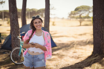 Teenage girl texting on smartphone at forest camp near tent and bicycle wearing sunglasses