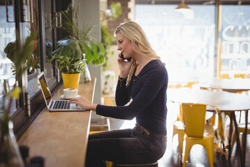 Woman juggling smartphone call and typing on laptop at cafe counter near coffee cup, copy space