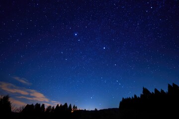 Naklejka premium A breathtaking nightscape featuring a vast, star-studded sky above a dark silhouette of a treeline on the horizon, with subtle hints of light possibly from a distant town or city