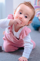 Cute baby daughter wearing pink overalls crawls on carpeted living room floor, exploring...