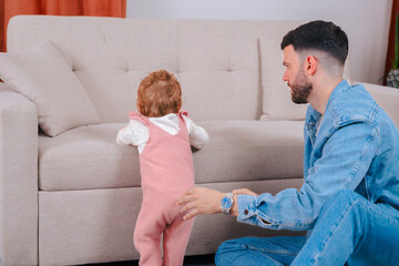 Loving father kneels on carpet supporting daughter in pink overalls as she practices standing up,...