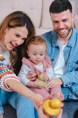 Smiling parents on living room sofa entertain their baby daughter in pink overalls with yellow duck...