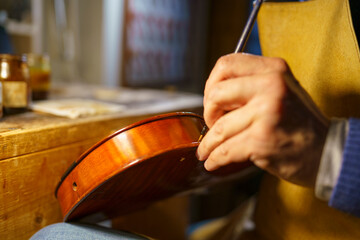 Violinmaker carefully varnishing wooden violin body, traditional tools and varnish jar surrounding workspace, highlighting intricate musical instrument crafting process