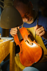 Skilled luthier inspecting handcrafted violin in wooden workshop, warm lamplight highlighting...