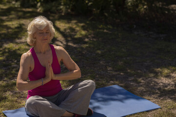 Senior woman wearing fitness bracelet sitting on blue yoga mat in park in dappled shade meditating