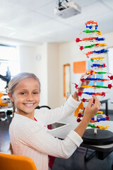 Girl assembling colorful DNA helix model at bright science lab near window with tablet, microscope