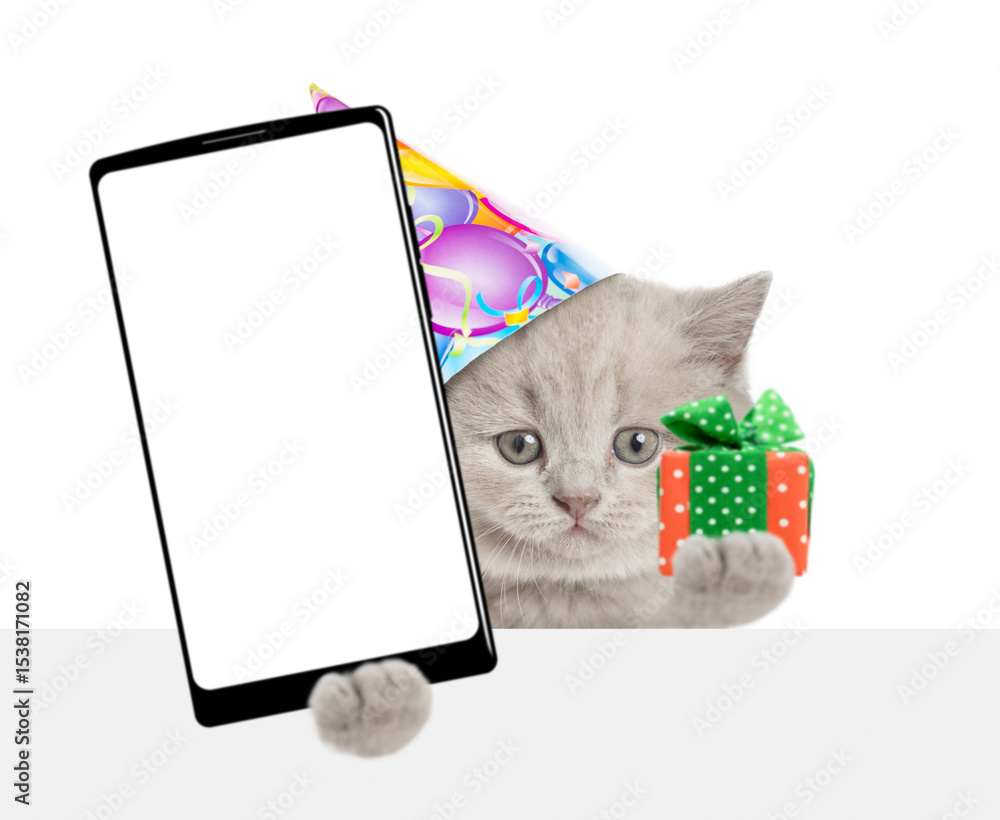 Poster cute tiny kitten wearing party cap shows gift box and big smartphone with blank screen above empty w