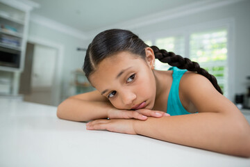 Girl leaning on white countertop by shuttered window in kitchen wearing turquoise top gazing out