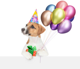 Funny jack russell terrier puppy wearing party cap holding balloons above blank white banner and looking at camera. isolated on white background